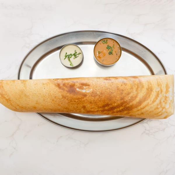 A golden-brown dosa, an Indian crepe, served with two small bowls of chutney and sambar on a steel plate.