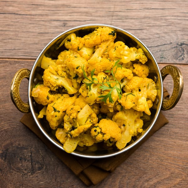 A brass bowl filled with vibrant yellow Indian Gobi Aloo, garnished with fresh cilantro, on a wooden table.