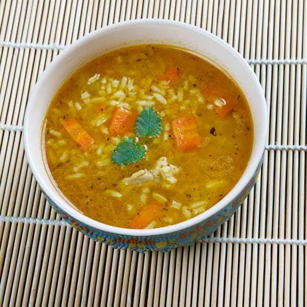 A bowl of Indian vegetarian soup with rice, carrots, and herbs on a bamboo mat.