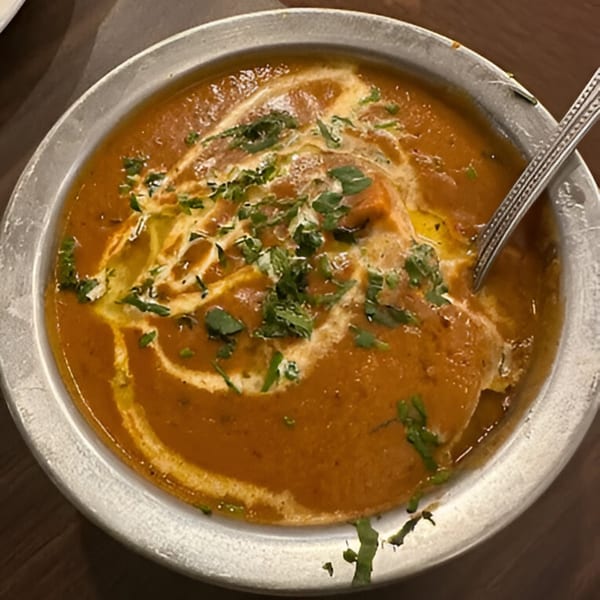 A close-up of a rich orange Indian curry, garnished with cream swirls and fresh cilantro, in a metal bowl.